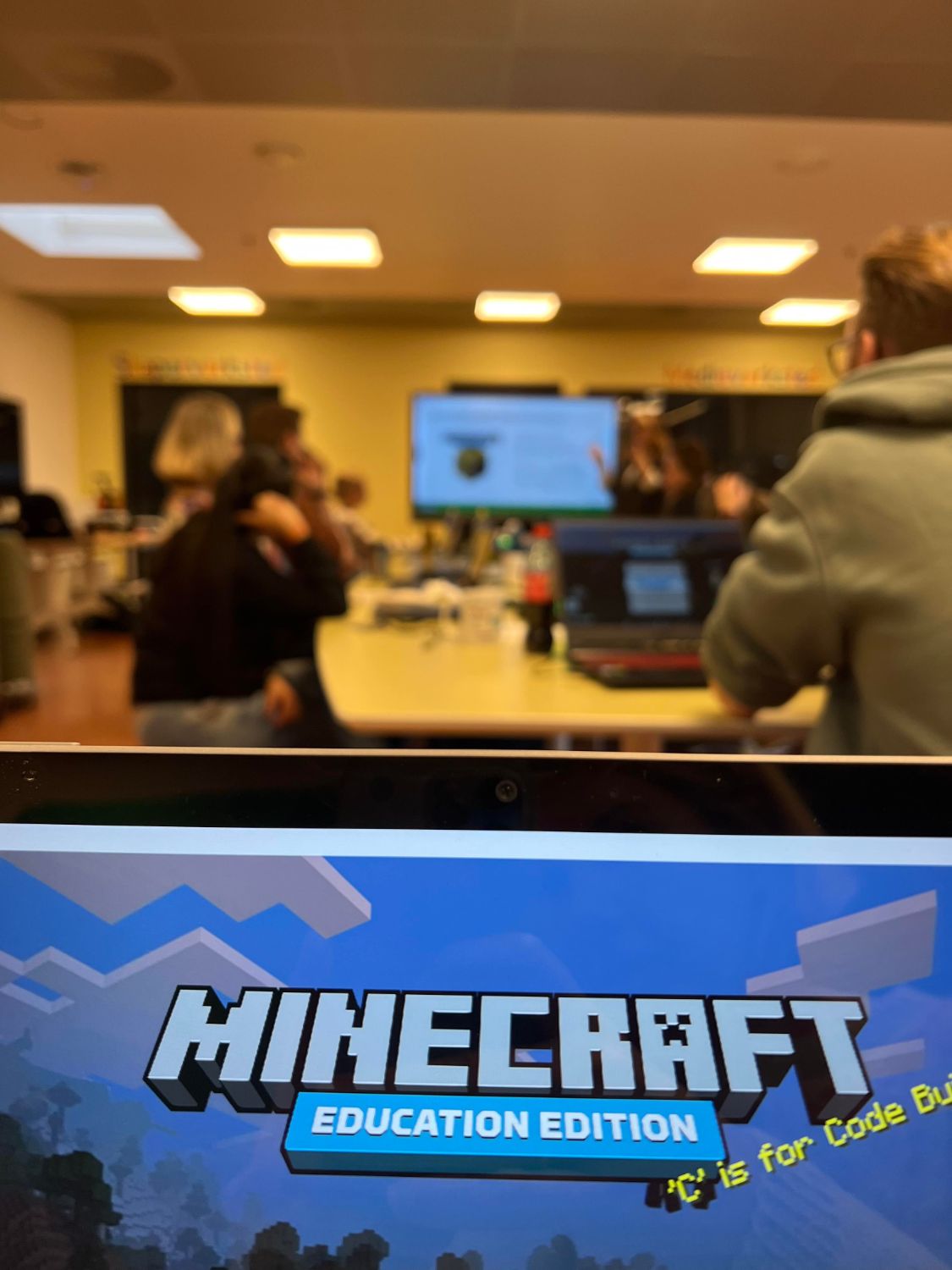 Viser PC med Minecraft logo og workshopdeltakere som sitter i bakgrunnen