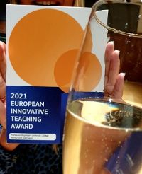 Her ses selve prisen, som er en plakett, for 2021 European Innovative Teaching Award. Et glass med Mozell holdes i forgrunnen. 