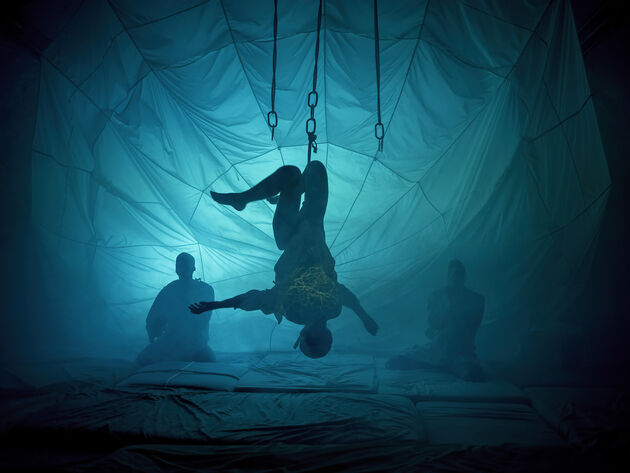 Foto på scene som viser en skikkelse som henger opp ned på scenen og en skikkelse sittende i bakgrunnen omgitt av et turkis lysskjær. 