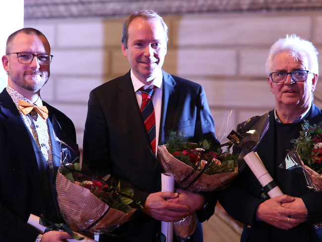 Bilde av de tre prisvinnerne ved siden av hverandre på scenen med blomster under arrangementet hvor prisen ble delt ut. 